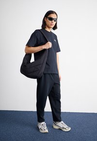 Schwarzes übergroßes T-Shirt mit Logo, schwarze gesteppte Umhängetasche, schwarze lässige Hose und sportliche weiße und graue Sneaker auf einem blauen Teppich.
