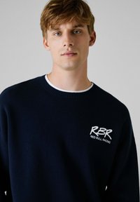 Granatowy sweter z miękkiego materiału, z okrągłym dekoltem i wyszywanym na biało logo "RBR Red Bull Racing" na piersi.