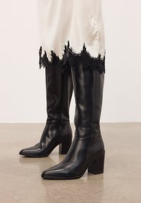 Bottes noires en cuir à hauteur de genou avec talons carrés, portées avec une robe crème ornée de dentelle noire, sur un fond de sol lisse.