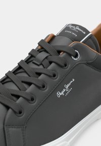 Κοντινή λήψη ενός μαύρου sneaker Pepe Jeans με μαύρες κορδόνες, λευκή σόλα και καφέ εσωτερική επένδυση, που δείχνει το λογότυπο στο πλάι και στην γλώσσα.