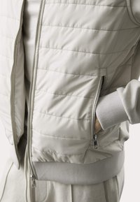 Gilet imbottito color beige chiaro con cuciture orizzontali, zip frontale e tasche laterali. Polsini e fondo a coste con texture liscia.