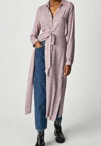 Robe longue à manches boutonnées en rose avec un motif géométrique, taille cintrée et fente sur le côté. Associée à un jean bleu taille haute et des bottines noires.