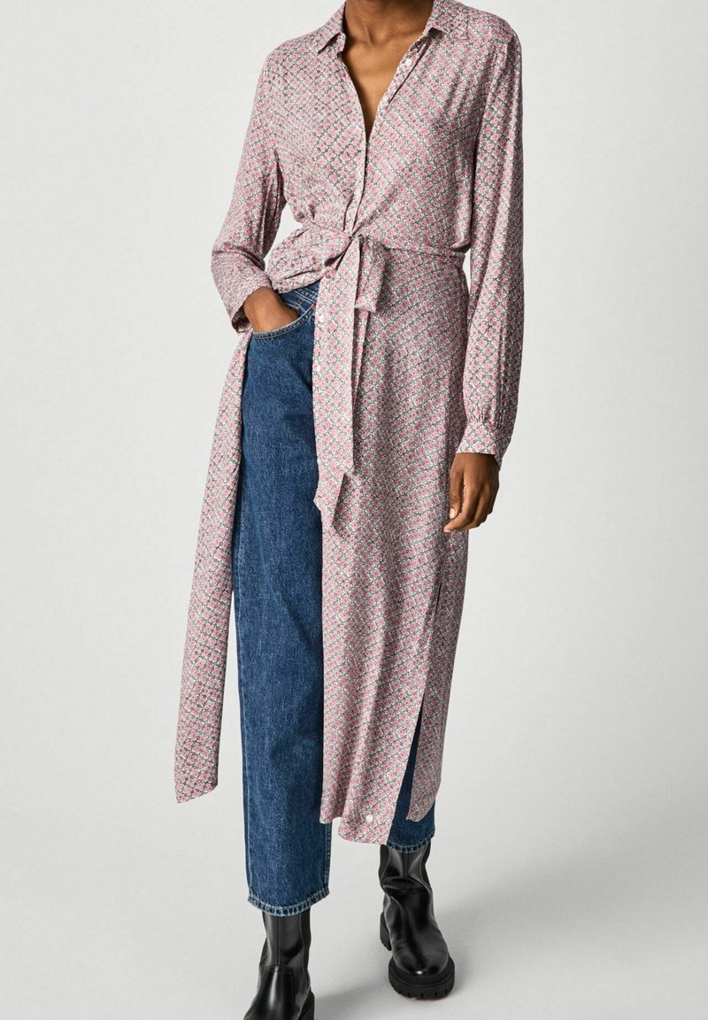 Robe longue à manches boutonnées en rose avec un motif géométrique, taille cintrée et fente sur le côté. Associée à un jean bleu taille haute et des bottines noires.