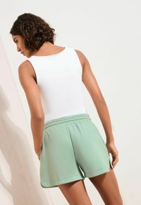 Top alb fără mâneci, asortat cu pantaloni scurți moi de culoare verde deschis, având o talie elastică și fante laterale lângă tiv.