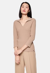 Junge Frau mit glattem, dunklem Haar trägt ein beiges, geripptes V-Ausschnitt-Top und passende weit geschnittene Hosen vor einfarbigem Hintergrund.