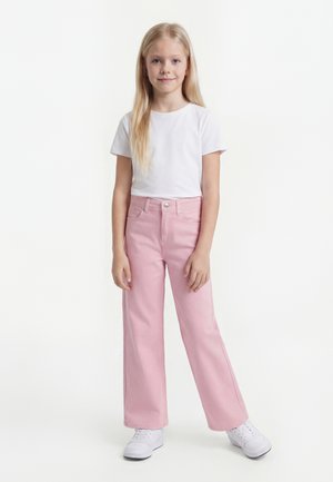 Jeune fille aux longs cheveux blonds portant un t-shirt blanc, un pantalon large rose et des baskets blanches, debout devant un fond blanc uni.