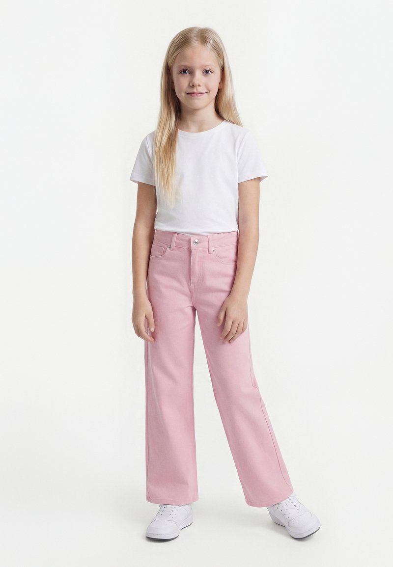 Jeune fille aux longs cheveux blonds portant un t-shirt blanc, un pantalon large rose et des baskets blanches, debout devant un fond blanc uni.