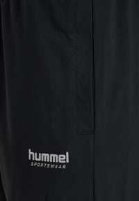 Närbild av svart sporttyg med en liten fick söm och den vita "hummel SPORTSWEAR" logotypen tryckt nära kanten.