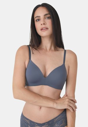 Frau mit langen dunklen Haaren, die einen grauen glatten BH und Spitzenunterwäsche trägt, steht mit einem Arm über dem Oberkörper verschränkt vor einem schlichten Hintergrund.