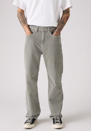 Persoon met getatoeëerde armen, gekleed in lichtgrijze corduroy broeken, een wit T-shirt en zwarte lage sneakers met witte veters, met de handen in de zakken.