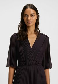 Tummanvioletti laskostettu mekko, jossa on V-pääntie, lyhyet läpikuultavat hihat ja teksturoidut yksityiskohdat vyötäröllä. Sileä, virtaava kangas ja jäsennelty malli.