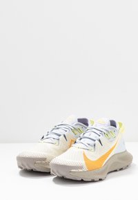 Chaussures de course avec une tige en mesh gris clair, des accents orange et jaune, et une semelle texturée de couleur blanc cassé. Dotées d'un système de laçage traditionnel.