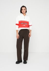 Λευκό και κόκκινο φούτερ με φερμουάρ και το γραφικό «Après Ski», σε συνδυασμό με καφέ παντελόνι φαρδιάς γραμμής. Διαθέτει άνετη εφαρμογή και μαύρα χοντρά παπούτσια.