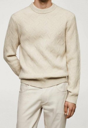 Mężczyzna w kremowym swetrze w warkocze z prążkowanym kołnierzem i mankietami, w zestawie z dopasowanymi kremowymi spodniami, stojący na jednolitym tle.