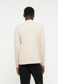 Camicia beige a maniche lunghe con scollatura a girocollo, in morbido tessuto misto cotone e polsini a coste, abbinata a jeans scuri. Design pulito e minimale.