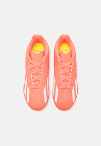 Scarpe da calcio corallo con strisce bianche, punta tonda e design con lacci. Presentano perforazioni per la traspirabilità e un sottopiede giallo.