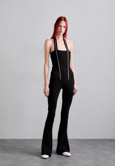 Black jumpsuit with a fitted bodice and flared legs, featuring adjustable straps and a textured fabric. White shoe accents complete the look.