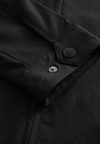 Detalle de una camisa negra de botones, con una textura suave, costuras de doble puntada y un detalle de botón a presión negro en el puño.