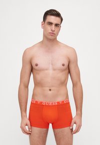 Boxeri scurți roșii din bumbac, cu un tiv elastic alb pe care este imprimat logo-ul "GUESS", design ajustat și textură netedă.