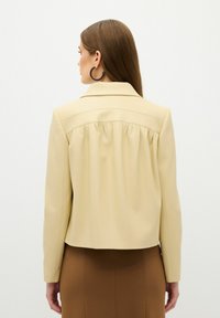 Beige Jacke mit geradem Schnitt, dezenten Falten hinten und Reverskragen. Getragen über einem braunen Rock.