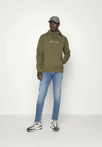 Mies, joka käyttää oliivinvihreää hupparia, jossa on teksti "TOMMY JEANS", vaaleansinisiä farkkuja, harmaata lippalakkia ja valkoisia sekä laivansinisä lenkkareita, seisoo valkoista taustaa vasten.