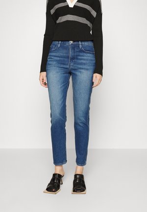 Vrouw met blauwe slim-fit jeans, zwarte gebreide trui met grijze strepen en zwarte open-toe sandalen met hak, staand tegen een witte achtergrond.