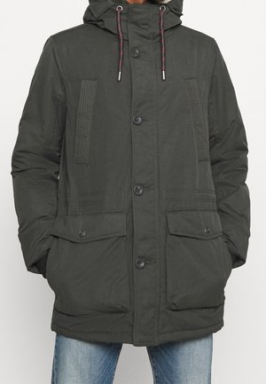 Dunkelgrüne, kapuzenbesetzte Winterjacke mit Knopfleiste, Kordelzügen, zwei Klappentaschen und seitlichen Eingrifftaschen, getragen über blauen Jeans.