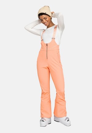 Peach-colored ski overalls with adjustable suspenders, zippered front, and side pockets. Worn over a long-sleeve white shirt and ski boots.