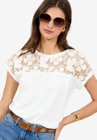 Haut blanc à manches courtes avec un empiècement en dentelle orné de motifs floraux. Porté avec un jean bleu taille haute et une ceinture marron. Grandes lunettes de soleil et bracelet en or.