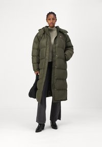 Maslinasto zelena puffer jakna s kapuljačom, kopčanjem na dugmad i zip, s bočnim džepovima, nosi se preko svijetlosive dukserice i crnih traperica.