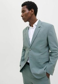 Costum verde deschis, croit cu reveri cu unghi drept, închidere cu un singur nasture și buzunare laterale. Stilizat cu o cămașă albă, textură fină și croială modernă.