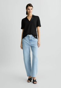 Zwarte polo met korte mouwen, een V-hals en textuurdetail, gecombineerd met lichtblauwe high-waisted jeans en zwarte open teen hakken.