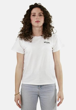 T-shirt bianca in cotone a maniche corte, con un piccolo logo nero "Moschino" sul petto. Abbinata a jeans azzurri chiari, a taglio dritto.
