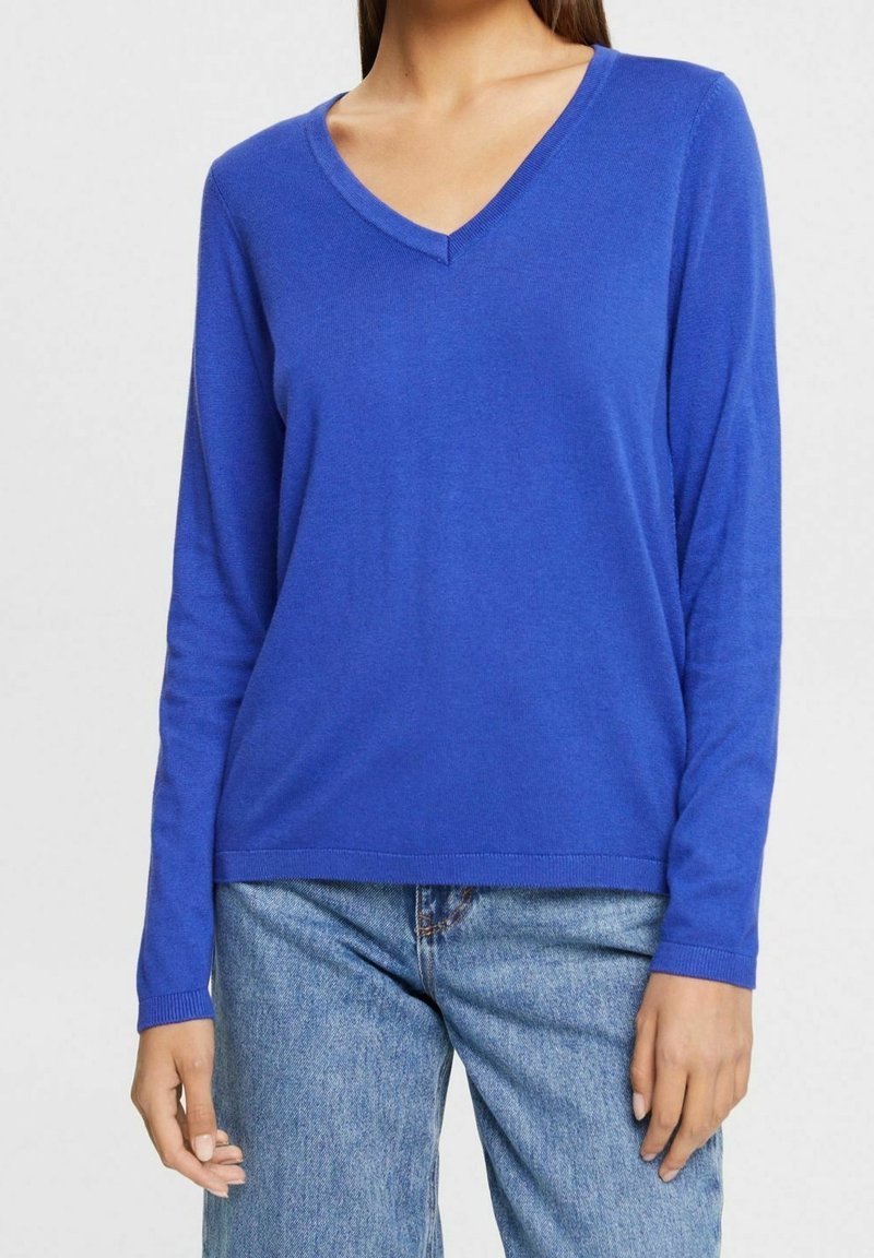 Femme portant un pull bleu uni à manches longues et col en V, ainsi qu'un jean en denim bleu clair, debout devant un fond uni.
