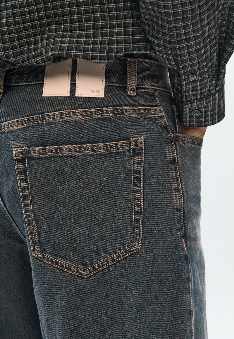 Dark denim jeans with a slightly distressed texture, featuring contrasting orange stitching and two rectangular leather labels on the waistband.