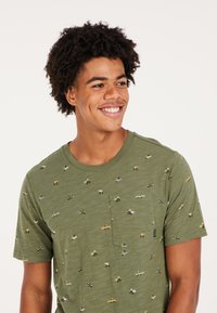 Žalia trumpų rankovių T-marškiniai su krūtinės kišene ir pakartotiniu mažų transporto priemonių ir banglenčių raštu. Minkšta tekstūra, laisvas fasonas.