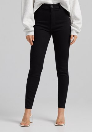 Femme portant un jean skinny noir, un sweat gris et des sandales à talons transparents, debout devant un fond uni gris clair.