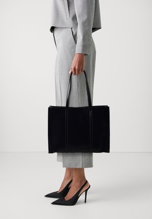 Femme portant un costume à carreaux noir et blanc, tenant un grand sac cabas noir, chaussée de talons aiguilles noirs, sur un fond uni.