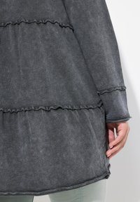 Dunkelgraues, geschichtetes Tunika mit ruffled Saum und Manschetten. Weicher, strukturierter Stoff, der Nahtdetails und eine lockere Passform aufweist.