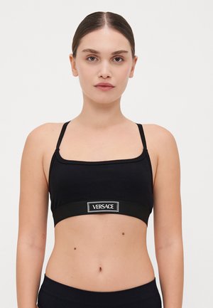 Melna ribota bralette ar regulējamiem siksnām, kura raksturīga plaša elastīga lente ar baltu "VERSACE" logotipu zīmola identitātei.