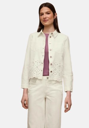 Giovane donna che indossa una giacca bianca con bottoni e motivo floreale traforato sopra una maglia malva e pantaloni bianchi abbinati.