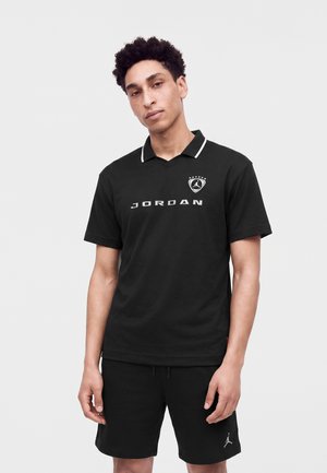 Νέος άνδρας με μαύρο πόλο μπλουζάκι Jordan με λογότυπο και μαύρα σορτς, στέκεται σε απλό φωτεινό φόντο.