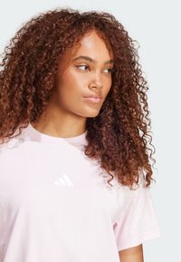 Tricou din bumbac roz deschis, cu o textură moale și mâneci scurte, având un logo alb Adidas pe piept.