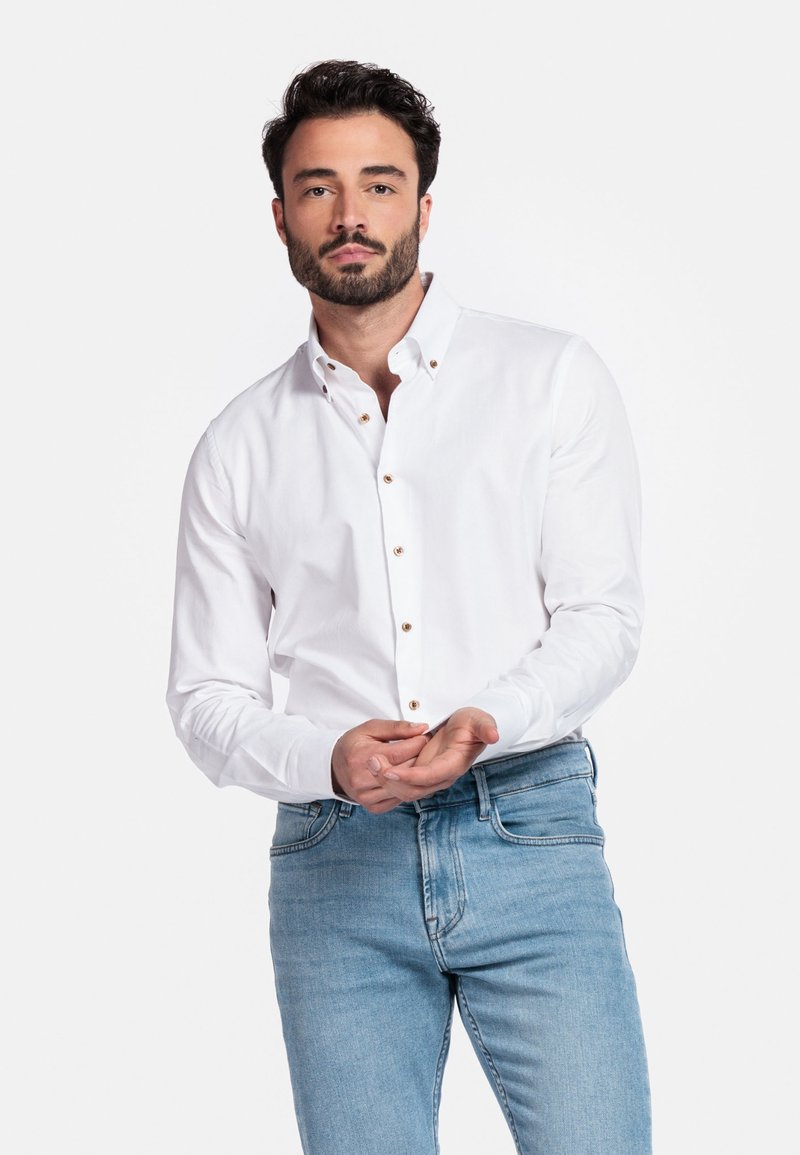 Witte knop-overhemd met een klassieke kraag, voorzien van zichtbare bruine knopen. G gedragen met lichtblauwe jeans, met de handen op de zoom van het shirt.
