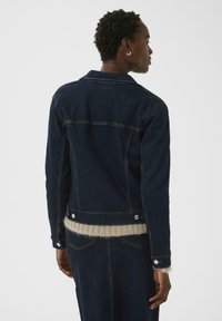 Dunkelblaue Jeansjacke mit Kragen, Rückenteil und silbernen Beschlägen. Sie hat einen figurbetonten Schnitt und gestickte Details und wird über einem leichten Pullover getragen.