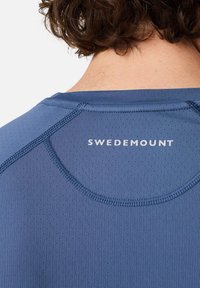 Das blaue Sportshirt besteht aus einem strukturierten Stoff mit kleinen Perforationen, hat einen abgerundeten Halsausschnitt und das auf der Rückseite aufgedruckte Logo "SWEDEMOUNT".
