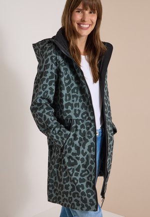 Frau trägt eine mittellange Kapuzenjacke mit schwarzem und dunkelgrünem Leopardenmuster, weißes Hemd und blaue Jeans, lächelnd mit den Händen in den Taschen.
