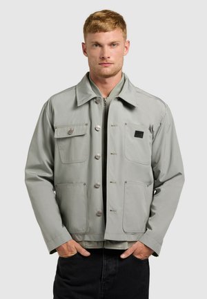 Fiatal férfi világosszürke, négy elejű zsebbel rendelkező gombos dzsekiben, kezét fekete nadrágzsebre téve, semleges háttér előtt áll.