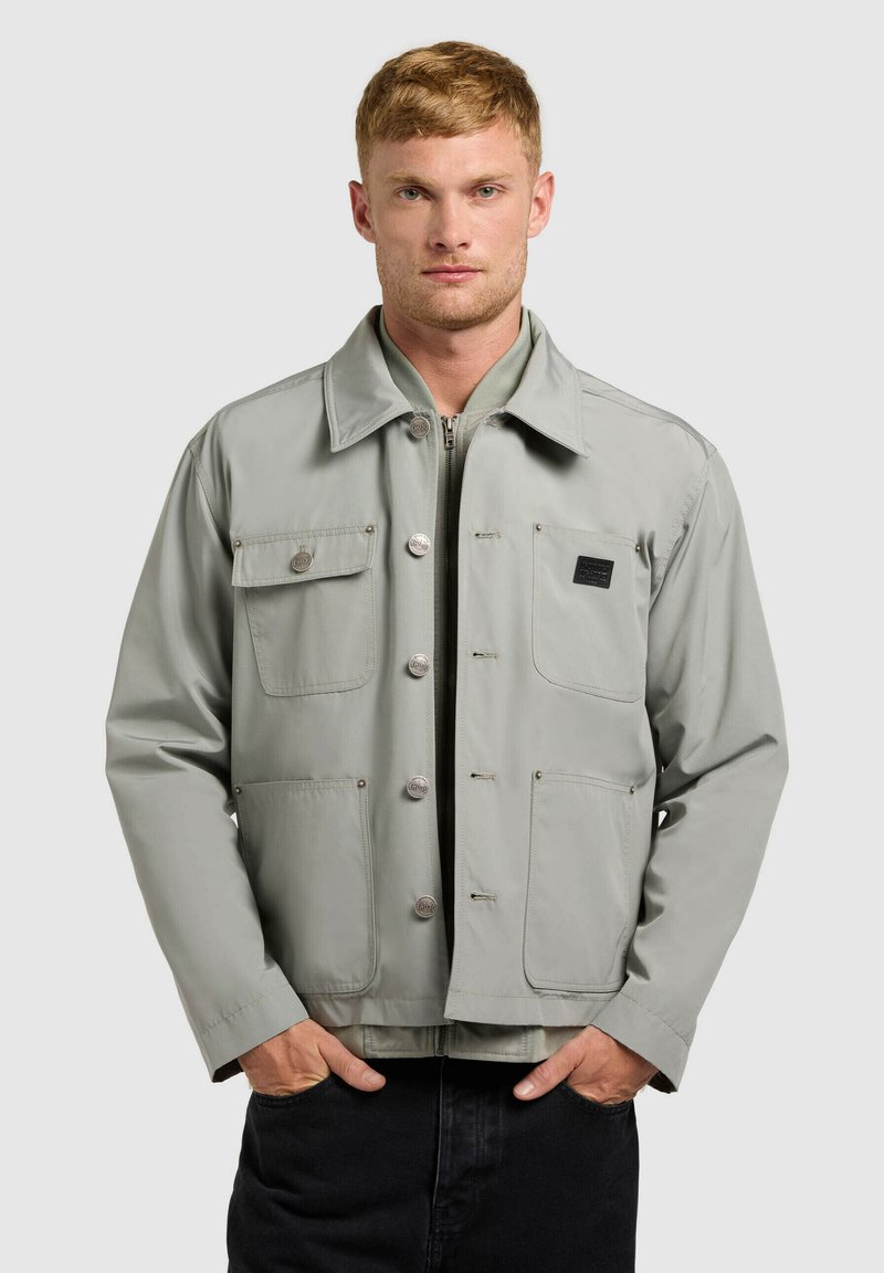 Jeune homme portant une veste grise claire boutonnée avec quatre poches avant, debout les mains dans les poches d'un pantalon noir, fond neutre.