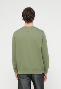 Άνδρας με καστανά σγουρά μαλλιά που φοράει ελιάς πράσινη μακρυμάνικη μπλούζα και σκούρο γκρι τζιν, στέκεται γυρίζοντας την πλάτη σε λευκό φόντο.